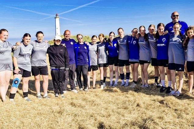 Gruppe von Sportlerinnen und Trainern in Sportkleidung stehen auf einer Wiese vor einem Leuchtturm unter blauem Himmel.