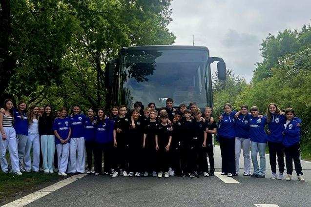 Gruppe junger Sportlerinnen in einheitlicher Kleidung posiert vor einem Bus auf einer Straße, umgeben von Bäumen.