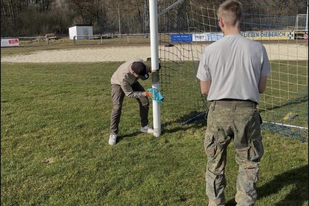 Zwei Personen befestigen ein Netz an einem Fußballtor auf einem Sportplatz bei sonnigem Wetter.